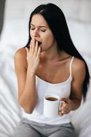Impossible to wake up Young beautiful woman with long dark hair holding a cup and yawning while sitting on the bed at home photo