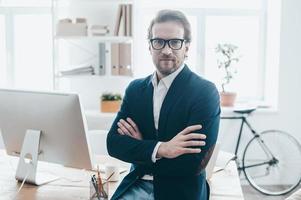 Welcome to my office. Handsome young man in eyeglasses keeping arms crossed and looking at camera while sitting on the table in creative office photo