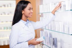 elegir el medicamento adecuado para usted. hermosa joven africana en bata de laboratorio eligiendo medicina mientras está de pie cerca de sí misma en la farmacia foto