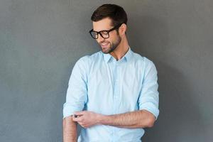 Preparing to work hard. Handsome young man in shirt adjusting sleeves and smiling while standing against grey background photo