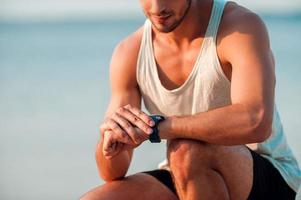 Going to beat his best time. Cropped image of young muscular man checking time on his watches while sitting outdoors photo