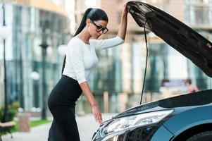 What is the problem here Side view of confident young businesswoman looking inside of the vehicle hood while standing near her car photo