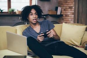 jugador salido. emocionado joven africano jugando videojuegos y manteniendo la boca abierta mientras se sienta en el sofá en casa foto