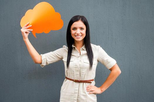 Put your thoughts in that cloud. Smiling young woman holding empty speech bubble and looking at camera while standing against grey background photo