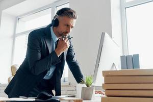 Thoughtful businessman in headphones looking at the computer while standing in office photo