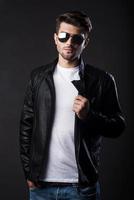 Trendy handsome. Handsome young man adjusting his leather jacket and looking at camera while standing against black background photo