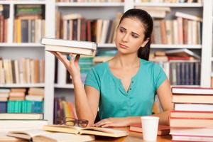 Tired student. Depressed young woman sitting at the library desk and holding a book stack in her hand photo