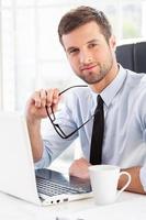 Making your business easier. Confident young man in formalwear holding glasses and looking at camera while sitting at his working place photo