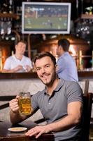 hombre en cervecería. apuesto joven sosteniendo una jarra de cerveza y sonriendo mientras está sentado en el bar foto