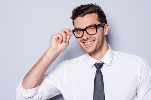 confiado y exitoso. retrato de un joven apuesto con camisa y corbata ajustándose los anteojos y mirando la cámara mientras se enfrenta a un fondo gris foto