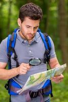 mapa de examen turístico confiado. joven confiado con mochila examinando el mapa con lupa mientras está de pie en la naturaleza foto