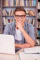 Smart and confident student. Confident young man holding hand on chin and smiling while sitting at the desk and in font of bookshelf photo