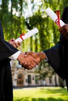 Congratulating with graduation. Close-up of two men in graduation gowns holding diplomas and shaking hands photo