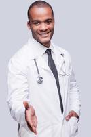 Always ready to help you. Cheerful African doctor stretching out hand for shaking while standing against grey background photo