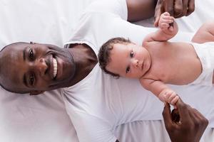 orgulloso de su bebé. vista superior de un joven africano feliz sosteniendo a su pequeño bebé y sonriendo mientras está acostado en la cama foto