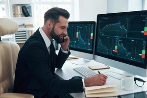 Concentrated young man in shirt and tie writing something down and talking on the phone while working in the office photo