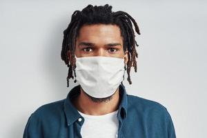 Young African man wearing medical face mask and looking at camera while standing against grey background photo