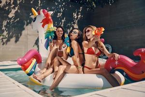 Attractive young women in swimwear smiling and having fun with squirt guns while floating on inflatable unicorn in swimming pool outdoors photo