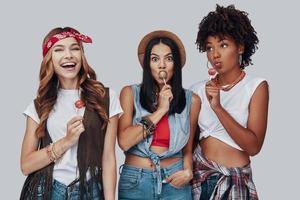 Three attractive young women eating lollipop and smiling while standing against grey background photo