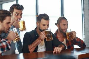 Group of handsome young men in casual clothing enjoying beer while sitting at the bar counter in pub photo