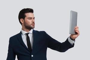 Hi Good looking young man in full suit holding digital tablet and looking at it while standing against grey background photo