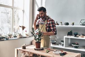 Taking order. Handsome young man in apron talking on the phone while standing in small garden center photo