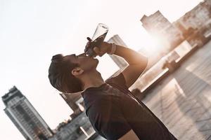 Hydrating. Young man in sports clothing drinking water while standing outside photo