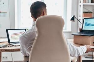 grandes datos vista trasera de un joven hombre de negocios con ropa formal analizando datos usando una computadora mientras está sentado en la oficina foto