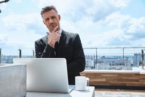 Confident mature man working on laptop and looking at camera while standing on the rooftop terrace photo