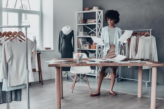 longitud completa de una joven africana mirando papeles y sonriendo mientras estaba de pie en su estudio cerca de la ropa colgada en los estantes foto