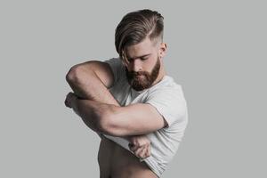 Too sexy for his clothes. Young man taking off his T-shirt while standing against grey background photo