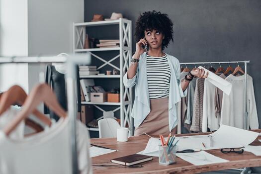 buscando la respuesta correcta. una joven africana pensativa hablando por teléfono inteligente y sosteniendo un papel mientras estaba en su taller foto