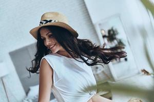 joven y hermoso. atractiva mujer joven con sombrero para el sol mirando hacia otro lado y sonriendo mientras está de pie en el dormitorio en casa foto
