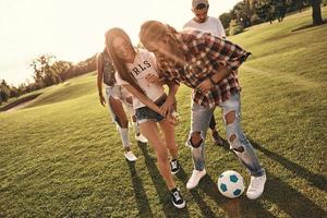 Full length of young smiling people in casual wear enjoying nice summer day while playing soccer outdoors photo