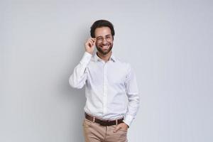 Handsome young man in white shirt looking at camera and adjusting his eyeglasses while standing against gray background photo