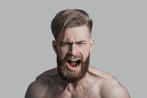 Feeling angry. Portrait of furious young man looking at camera and shouting while being in front of grey background photo