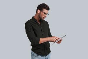 Examining his digital tablet. Serious young man in eyeglasses working on his digital tablet while standing against grey background photo