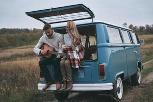 pareja romantica. un joven apuesto tocando la guitarra para su bella novia mientras se sienta en el maletero de una mini furgoneta de estilo retro azul foto