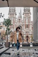 día increíble en la ciudad. longitud completa de una joven atractiva mirando hacia otro lado y sosteniendo una taza desechable mientras está de pie al aire libre con la iglesia en el fondo foto