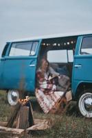 involucrados en la lectura. atractiva mujer joven cubierta con una manta leyendo un libro mientras se sienta dentro de la mini furgoneta de estilo retro azul foto