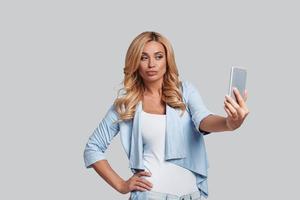 hermosa autofoto atractiva mujer joven con la mano en la cadera y tomando selfie mientras está de pie contra el fondo gris foto