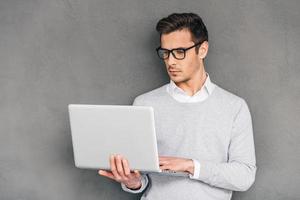 Checking his timetable. Confident young man holding laptop and typing while standing against grey background photo