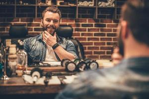 Enjoying his perfect beard. Handsome young bearded man looking at his reflection in the mirror and keeping hand on chin while sitting in chair at barbershop photo