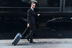 Confident businessman. Full length of young man in full suit pulling luggage and checking the time while walking outdoors photo