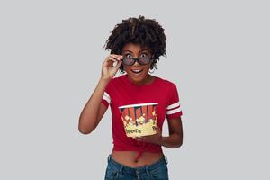 Attractive young African woman in 3D eyewear eating popcorn and looking at camera while standing against grey background photo