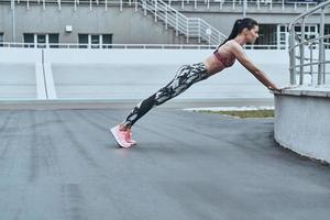 Never give up. Beautiful young woman in sports clothing keeping plank position while exercising outdoors photo