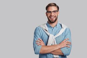 Confident in his style.  Good looking young man keeping arms crossed and looking at camera with smile while standing against grey background photo