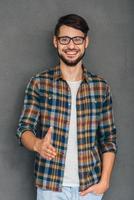 Nice to meet you Cheerful young man reaching out his hand in greeting you and holding hand in pocket while standing against grey background photo