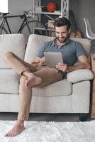 Enjoying his free time at home. Handsome young man using his digital tablet with smile while sitting on the couch at home photo