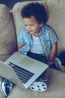 I like this laptop. High angle view of little African baby boy looking at laptop while sitting on the couch at home photo